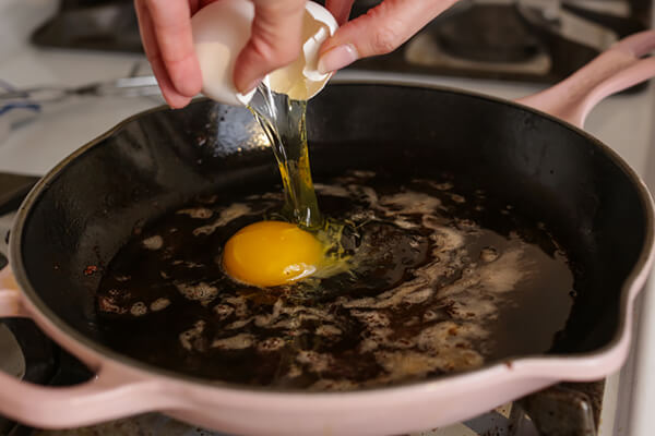 Hand cracking an egg into a skillet full of bacon grease.